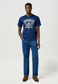 T-shirt in cotone blu navy con la scritta "WRANGLER 1947" stampata in bianco, abbinata a jeans blu e stivali marroni. Design semplice con scollatura a girocollo.