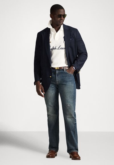 Moški z očali za sonce, mornarsko modrim blazerjem, belo polo majico z napisom, modrimi kavbojkami, rjavim pasom in rjavimi usnjenimi čevlji, stoji z roko v žepu.