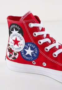 Sneaker alta in tela rossa con lacci bianchi, caratterizzata da diversi marchi circolari in nero, bianco e blu sul lato.