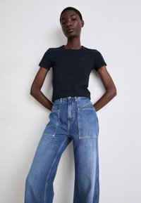 Tricou ajustat de culoare bleumarin, cu mâneci scurte, asortat cu blugi largi de culoare albastră deschisă, cu talie înaltă, având buzunare frontale și detalii vizibile de cusătură.