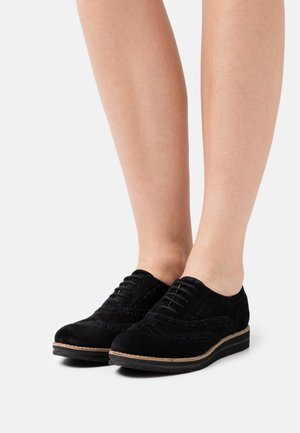 Chaussures brogue en suède noir à lacets avec semelle contrastante portées pieds nus sur fond blanc.