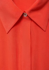 Camicia rossa intrecciata con colletto a punta, chiusura con un solo bottone e una texture liscia con dettaglio plissettato lungo il centro.