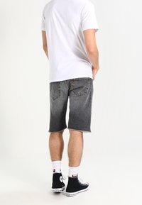 Hombre vestido con una camiseta blanca, pantalones cortos de mezclilla negros hasta la rodilla, calcetines blancos y zapatillas altas negras, de pie con las manos en los bolsillos.