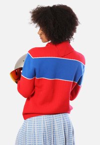 Maglione lavorato a maglia rosso e blu con colletto alto e texture a coste. Presenta un design a blocchi di colore orizzontali ed è abbinato a una gonna blu con motivo.