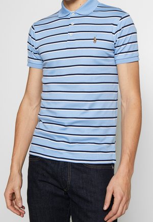 Homme portant un polo bleu clair à rayures horizontales foncées et un jean foncé, debout devant un fond uni.