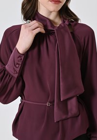 Blusa bordeaux con dettaglio a fiocco sul colletto, maniche lunghe con rifiniture a bottone e vita con cintura. Tessuto liscio e texture interessante.