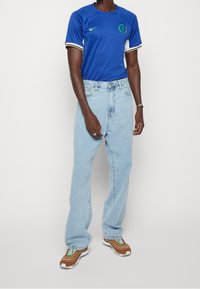 Camiseta de fútbol azul de Nike con logo verde, mangas cortas y rayas en blanco y negro. Pantalones vaqueros holgados de color azul claro y zapatillas deportivas marrones.