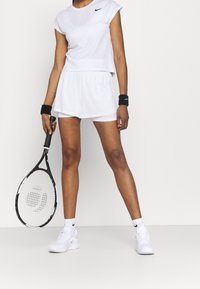 Conjunto atlético blanco compuesto por una camiseta ajustada y pantalones cortos, hecho de tela ligera. Logo de Nike en negro, combinado con zapatillas blancas y muñequeras.