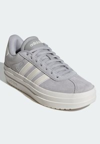 Grå mockaskor med rund tå, tre vita ränder på sidan, och vadderad krage. Vit gummisul. Minimalistisk design.