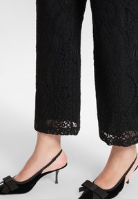 Czarne koronkowe spodnie o skomplikowanym wzorze, zestawione z czarnymi sandałami na obcasie typu slingback z kokardą i wąskim obcasem.