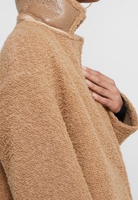 Abrigo texturizado beige con una apariencia suave y peluda, y acentos marrones brillantes en el cuello. El diseño es de gran tamaño, con un ajuste relajado.