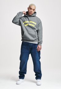 Felpa grigia con scritta gialla "F*ck Your Bad Vibes". Abbinata a jeans blu scuro e sneakers bianche. Il modello si trova in un ambiente neutro.