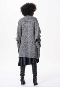 Grosse cardigan en tricot gris avec épaules tombantes, poignets côtelés et une étiquette en tissu dans le dos. Assorti avec une jupe noire et des bottes noires hautes.