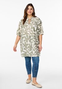 Tunika leggera con motivo floreale in verde e beige, con scollatura a oblò e laccetto, maniche a palloncino corte e vestibilità comoda, abbinata a jeans blu.