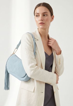 Femme en blazer beige et robe gris foncé porte un sac à bandoulière bleu clair matelassé avec un pompon, regardant de côté sur fond uni.
