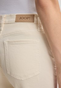 Jeans in denim beige con una tasca posteriore visibile. Etichetta in pelle con "JOOP!" in marrone attaccata alla vita. Texture liscia e design aderente.