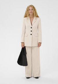 Costume beige sur mesure avec un blazer cintré présentant trois boutons noirs et deux poches. Pantalon à jambes larges et sac à main noir. Texture lisse.