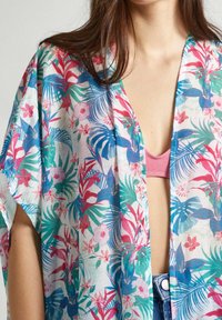 Kvinna som bär en färgglad blommig kimono över en rosa topp och blå jeans, med rakt brunt hår.