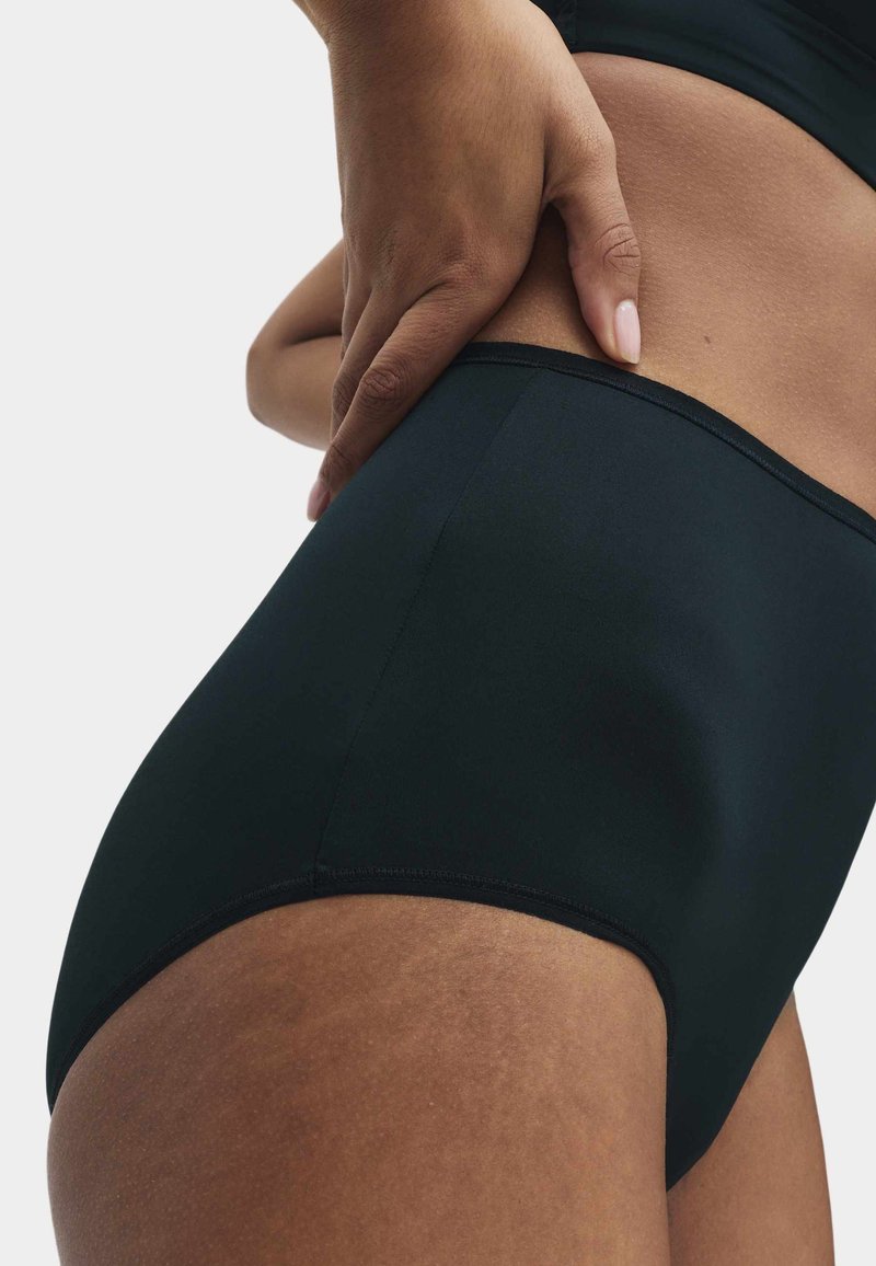 Close-up of a person wearing high-waisted black underwear with one hand resting on their hip against a plain background.