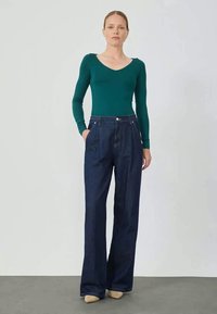 Bodysuit ajustado de mangas longas verde, com decote em V, combinado com jeans de perna larga de cintura alta em azul escuro. Os jeans apresentam bolsos frontais e fecho com botão.