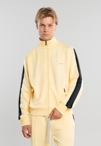 Hombre rubio con chándal amarillo claro con rayas negras en las mangas, de pie contra un fondo liso gris claro.