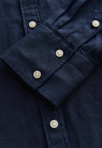 Chemise bleu marine boutonnée avec un tissu texturé, des poignets doubles avec quatre boutons blancs, et un design épuré et classique.