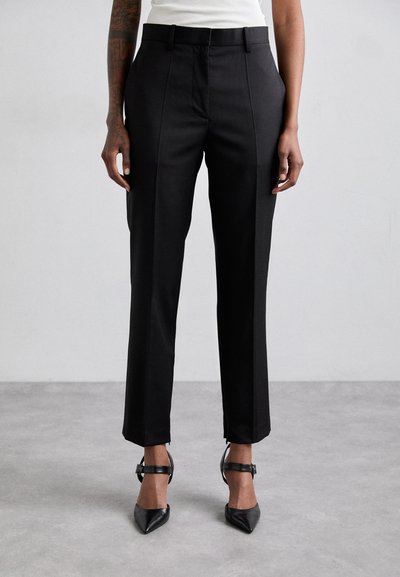 Pantalones negros ajustados con corte recto, con un frente plano, trabillas para el cinturón y costuras sutiles a lo largo del costado. Combinados con tacones negros puntiagudos.