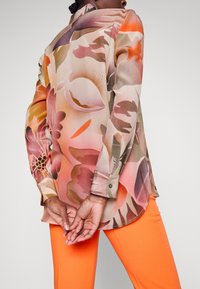 Blouse à manches longues en tissu transparent avec un motif floral coloré dans des tons de rose, beige et orange. Comprend des poignets boutonnés et un ourlet arrondi.