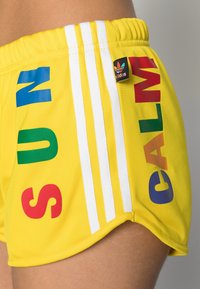 Žluté sportovní šortky s barevným nápisem "SUN" a "CALM" v červené a modré barvě. Mají bílé postranní pruhy a malou nášivku s logem Adidas.