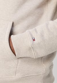 Sudadera beige con puños y dobladillo acanalados, que presenta un bolsillo tipo canguro en la parte frontal. Pequeño parche de logo en rojo, blanco y azul en el puño izquierdo.