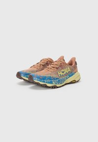 Trailrunning-Schuhe mit einem rosa Mesh-Obermaterial, blauen gemusterten Zwischensohlen und gelben Akzenten. Sie verfügen über eine robuste braune Sohle für Traktion.