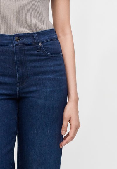 Gros plan d'une personne portant un jean bleu foncé et un top sans manches gris clair, montrant la taille et le bras droit sur un fond uni.