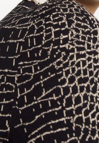 Gros plan sur un tissu noir avec un motif de grille blanche irrégulière porté sur une épaule, montrant une partie d’un cou et une boucle d’oreille en perle.