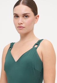 Maillot de bain vert avec un design en V, featuring des bretelles ajustables avec des accents en hardware rectangulaires et une texture de tissu douce et lisse.