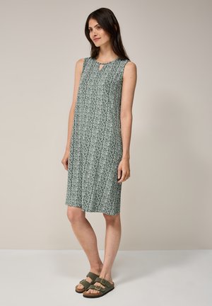 Ärmelloses grünes Kleid mit einem durchgängigen geometrischen Muster, einem Schlitzdetail am Ausschnitt und einer lockeren Passform, kombiniert mit olivfarbenen Sandalen.
