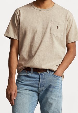 Mann trägt beige T-Shirt mit kurzen Ärmeln und Brusttasche, versehen mit kleinem gesticktem Logo, und hellblaue Jeans, lässig die Hand in der Tasche.