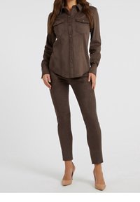 Vrouw gekleed in een nauwsluitend bruine suède overhemd met knopen, flapzakken en bijpassende slanke broek, gecombineerd met beige hoge hakken.