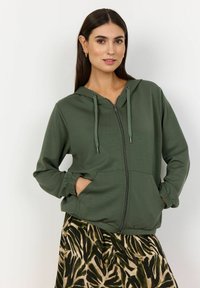 Zelená mikina na zips s kapucňou na šnúrku, elastickými manžetami a prednými vreckami, skombinovaná s patterned sukňou v prírodných odtieňoch.