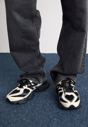Personne portant des chaussures de sport noir et blanc avec des lacets noirs et un jean gris foncé, debout sur un tapis bleu.