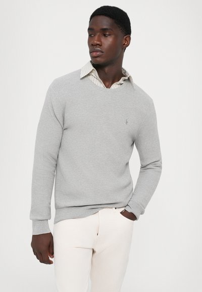 Suéter gris de punto con cuello redondo, puños acanalados y un pequeño logo. Lleva encima una camisa de cuello con rayas claras. Modelo con pantalones neutros.