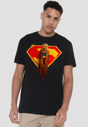 Junger Mann trägt ein schwarzes T-Shirt mit rotem und gelbem Superman-Logo und einer Figur in braunem Mantel auf der Vorderseite.