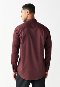 Camicia a maniche lunghe color bordeaux con texture liscia, colletto a punta e polsini con bottoni, con orlo curvo e sottili dettagli di cucitura sul retro.