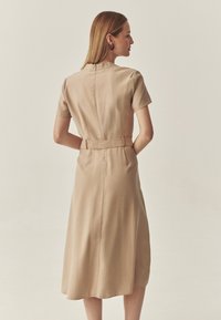 Beige Kleid in Kniehöhe mit kurzen Ärmeln, einem taillierten Oberteil und einem Gürtel in der Taille. Es ist aus einem glatten, fließenden Stoff gefertigt und hat einen diskreten Reißverschluss auf der Rückseite.