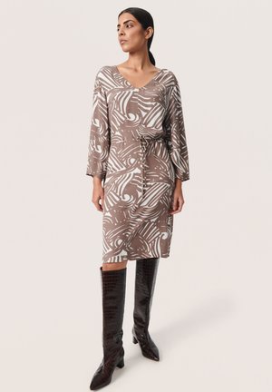Robe à motifs marron et blanc avec un col en V, des manches longues et une taille à cordon, associée à des bottes brillantes marron foncé jusqu'aux genoux.