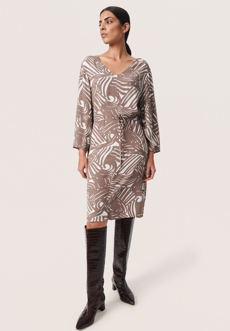 Robe à motifs marron et blanc avec un col en V, des manches longues et une taille à cordon, associée à des bottes brillantes marron foncé jusqu'aux genoux.