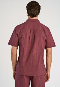 Camicia a maniche corte in tessuto bordeaux strutturato, con colletto, orlo dritto e vestibilità rilassata. Nessuna hardware o motivo visibile.
