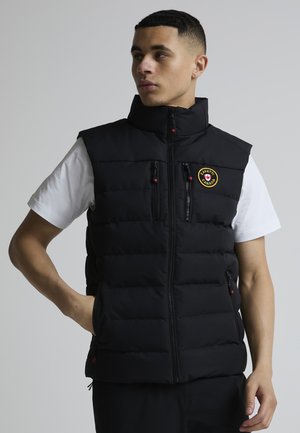 Ung mann med svart vattert vest med glidelås foran og en gul rund lapp, over en hvit kortermet skjorte, ser mot venstre.