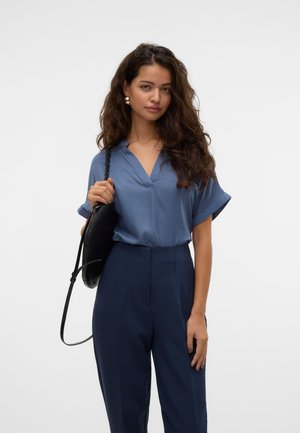 Mujer con cabello largo y rizado, vistiendo una blusa azul de manga corta y pantalones de talle alto en color azul marino, sosteniendo un bolso negro de hombro sobre un fondo blanco.