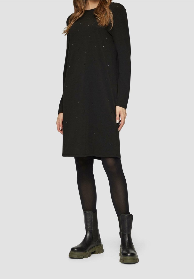 Femme portant une robe noire à manches longues, longueur genoux, avec des petits pois, des collants noirs et des bottes noires épaisses avec des semelles vertes.