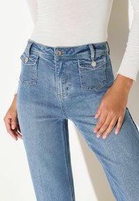 Hellblaue Jeans aus Denim mit hoher Taille, ausgestattet mit zwei Vorder- und zwei Gesäßtaschen, akzentuiert durch silberne Knopfbeschläge.